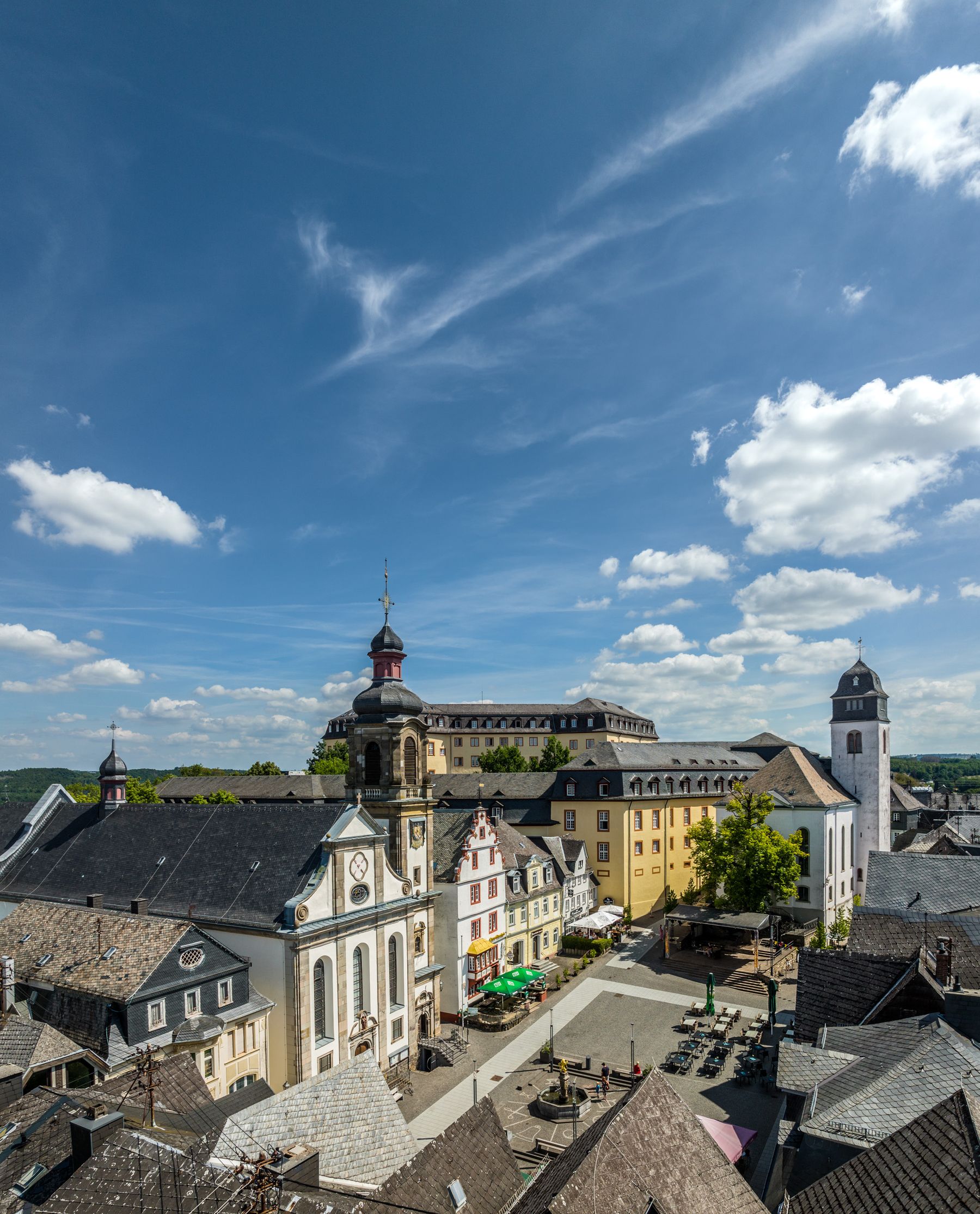 Eine malerische Ansicht eines europäischen Stadtplatzes mit historischen Gebäuden, darunter eine Kirche mit einem Uhrenturm und ein großes gelbes Gebäude, unter einem strahlend blauen Himmel mit vereinzelten Wolken.